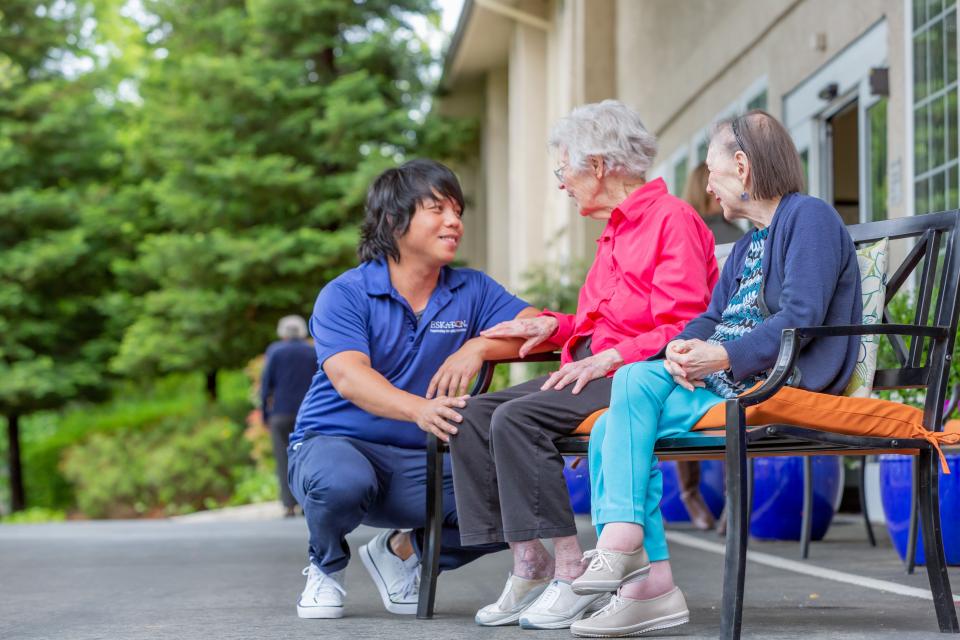 Two women resident sitting outside on a bench smiling and talking with a staff member.